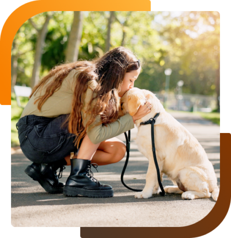 A woman crouches in a park, gently holding and comforting a seated golden retriever on a leash.